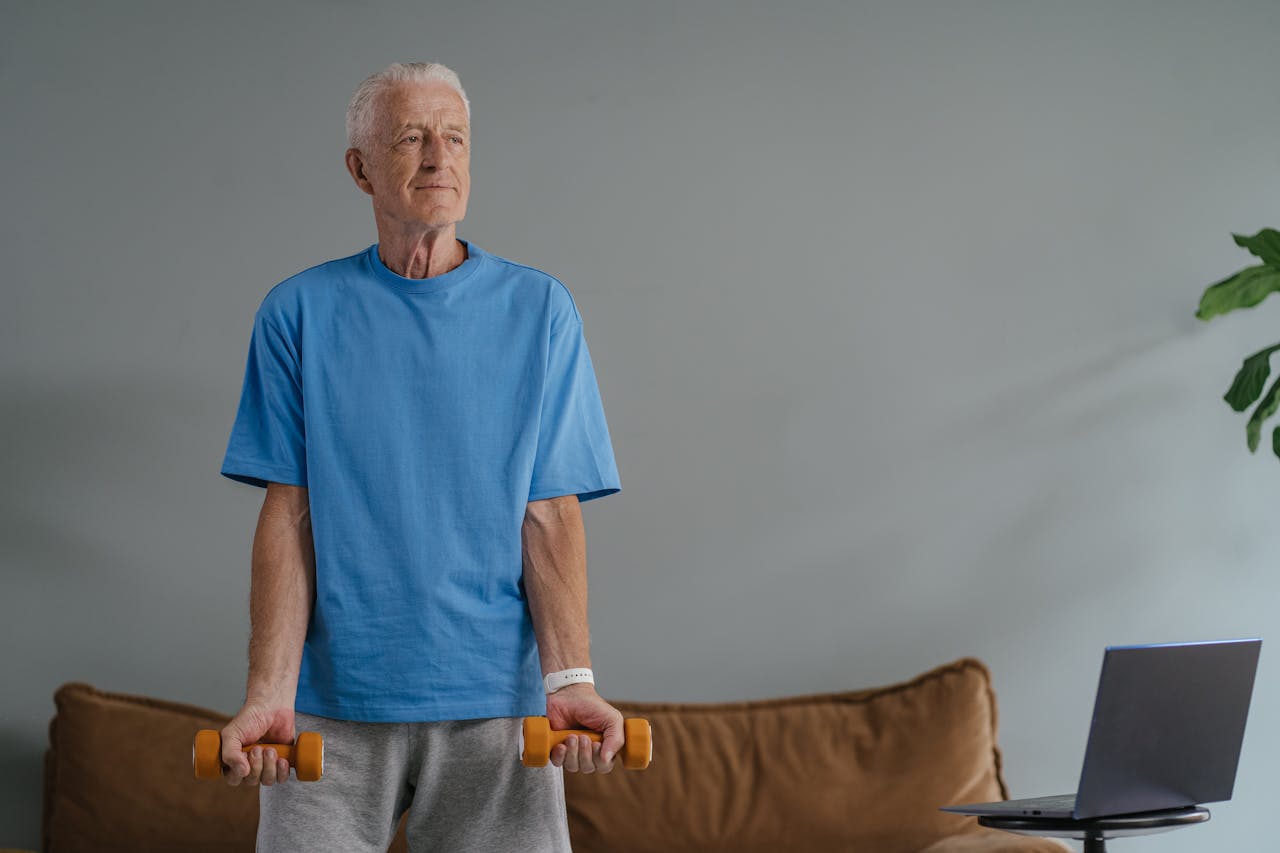 about-01 Elderly man in blue shirt lifting dumbbells near laptop, embracing home fitness.